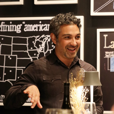 Man in a dark shirt standing in front of a blackboard with text and a map, holding a bottle.
