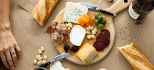 Image of a cheese plate with three cheeses and accompaniments including wine and baguette