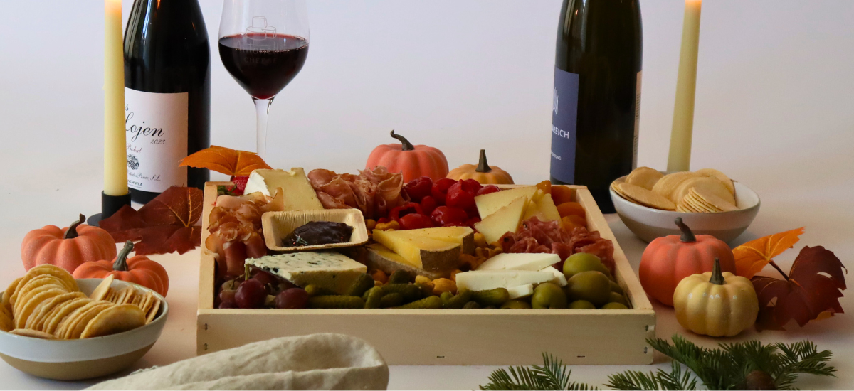 Cheeseboard with assorted cheeses, fruits, and crackers on a wooden board, accompanied by wine bottles and glasses.
